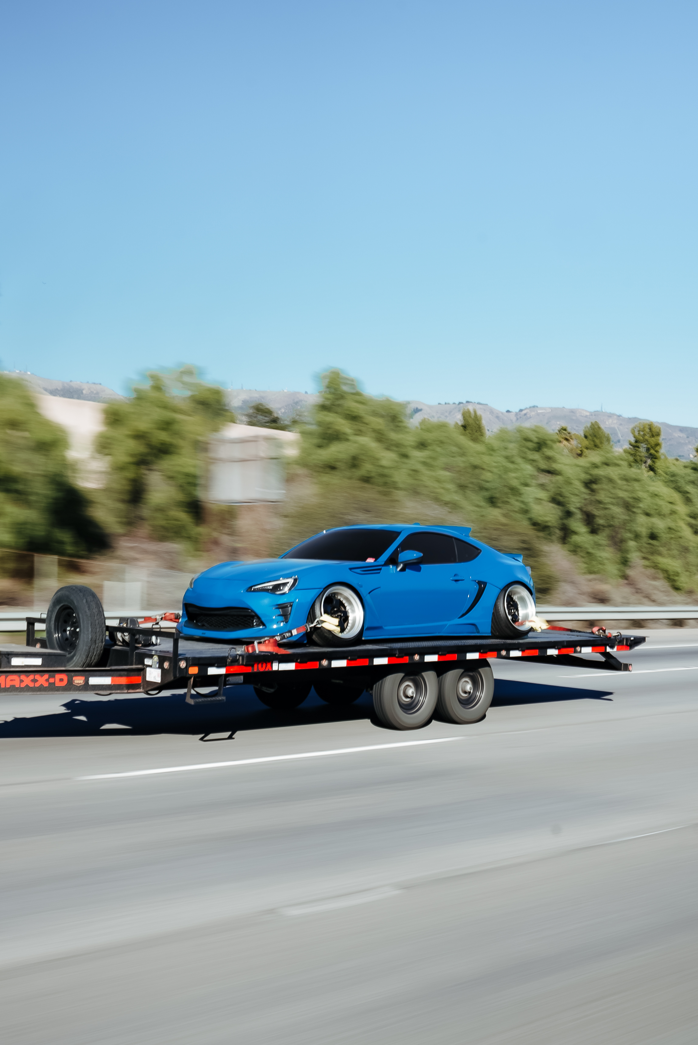 Dark gray sports car with modified wheels on black trailer attached to white pickup truck at sunset.