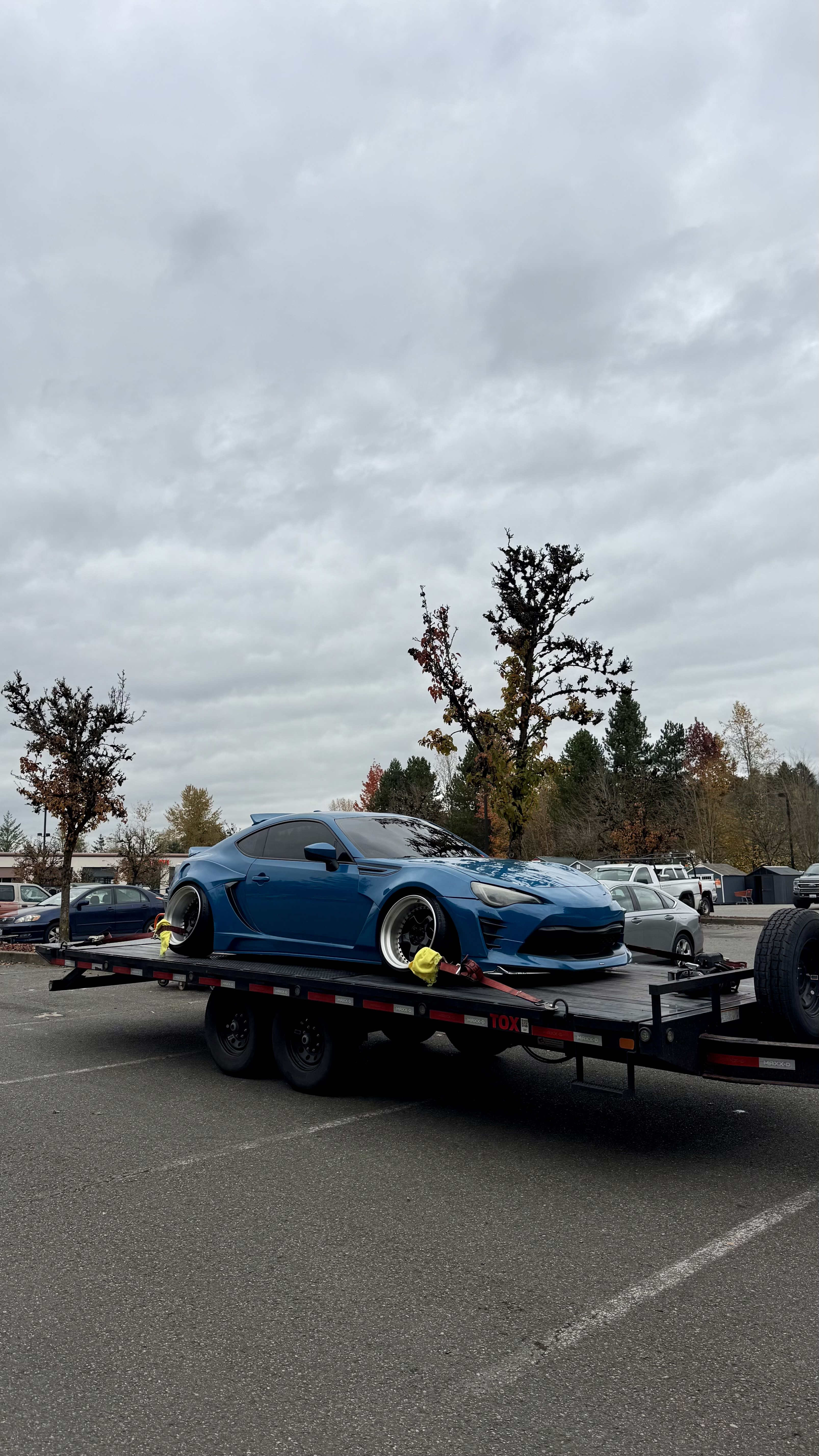 Dark sedan with lowered wheels on an open car trailer attached to a white pickup truck at sunset.