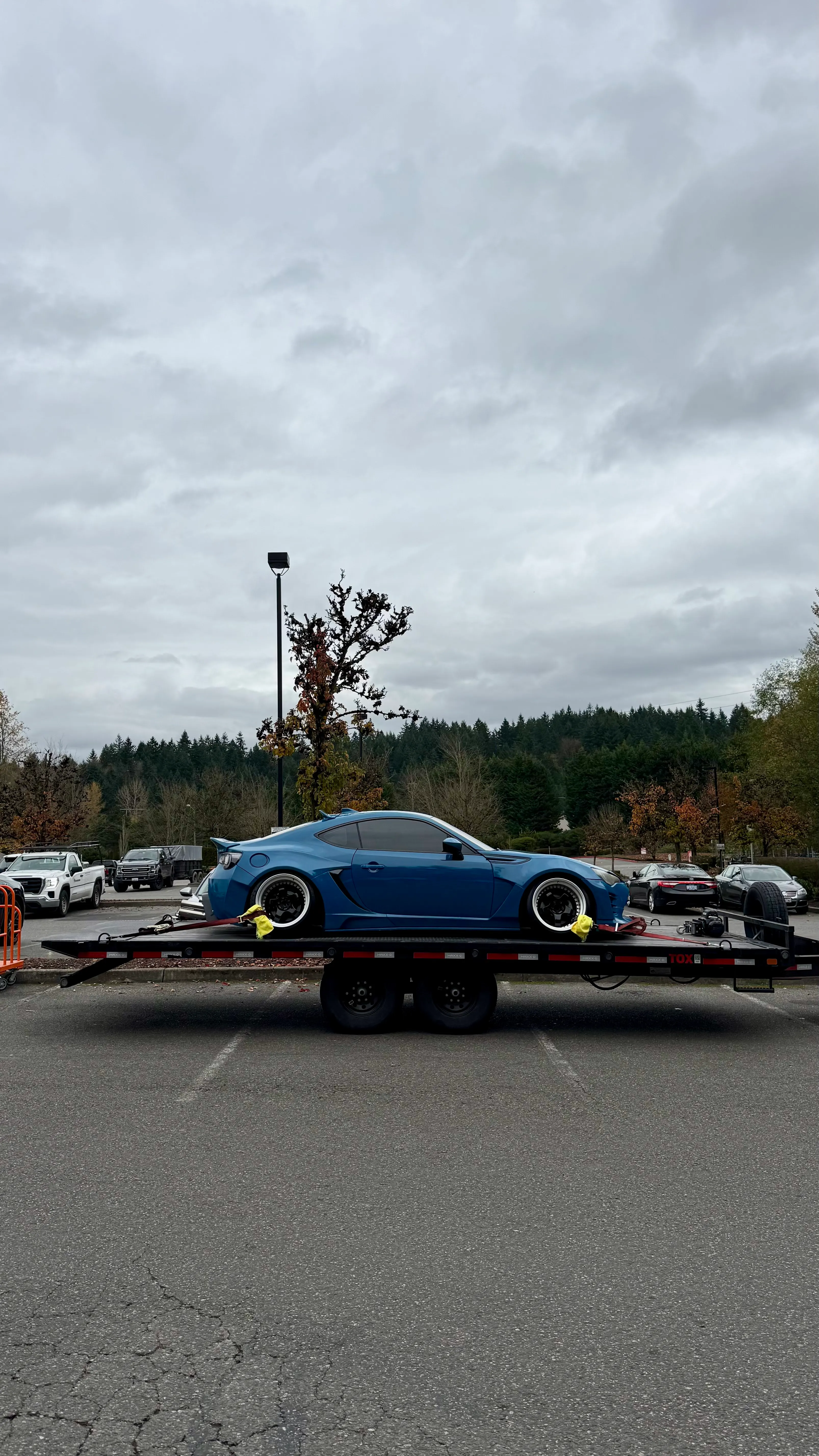 Blue sports car with wide wheels secured on a flatbed trailer in a parking lot with trees and cars behind.