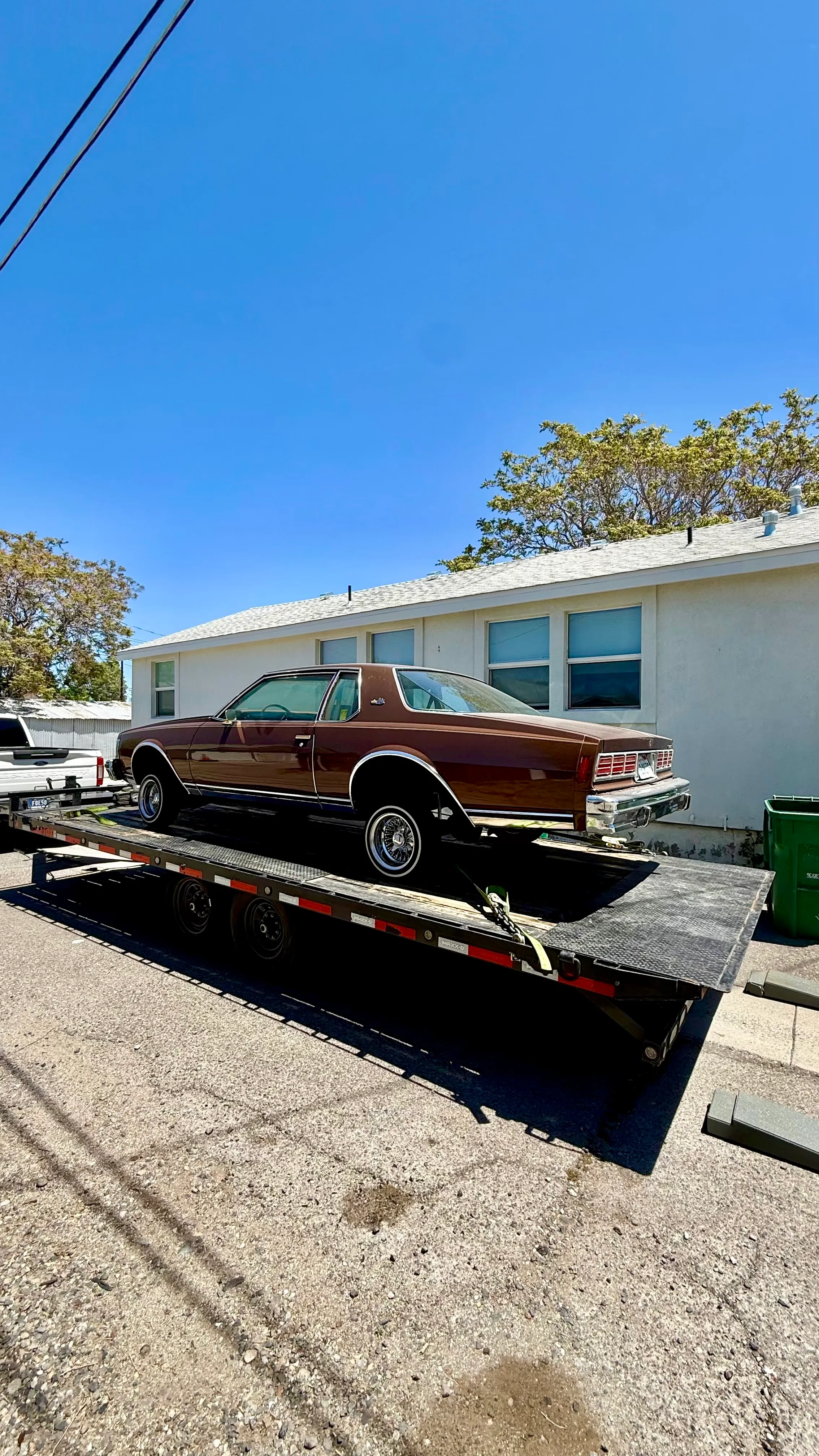 Brown classic car with raised rear wheels on black flatbed trailer parked by a white building.