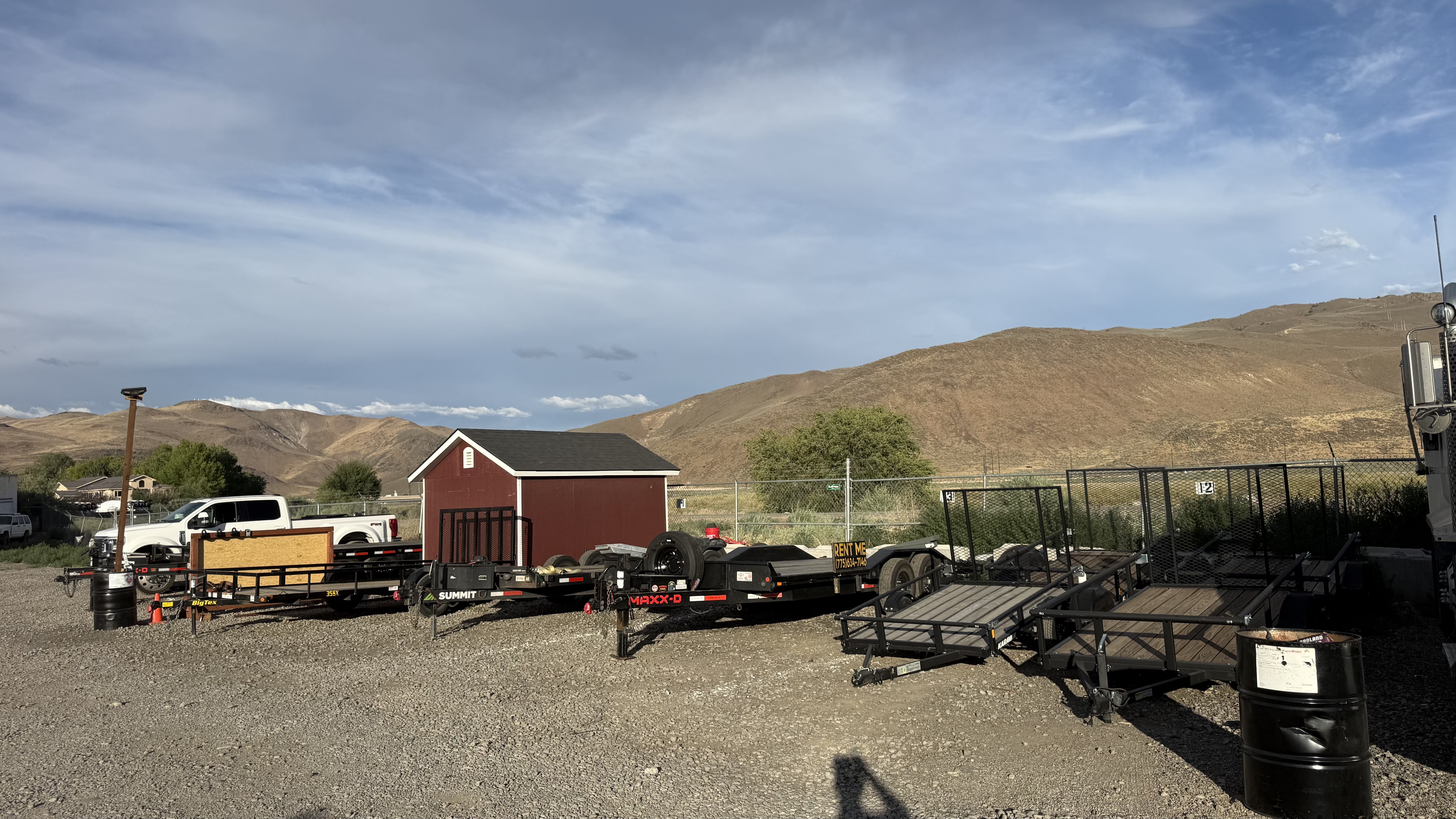Outdoor lot with various flatbed trailers and a small red shed by dry hills under a partly cloudy sky.