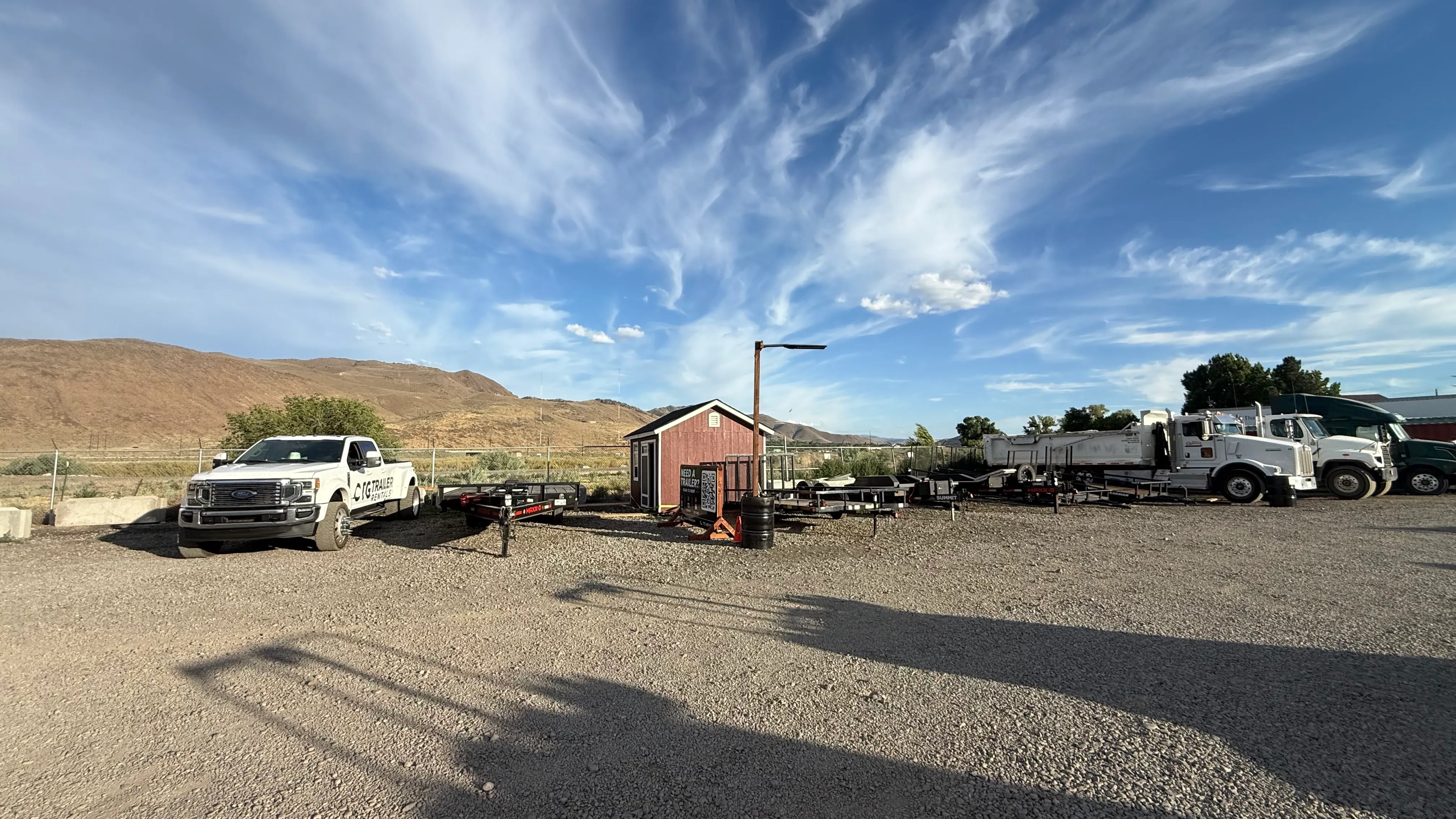 Gravel lot with white trucks, utility trailers, a small shed, and mountains under a blue sky.