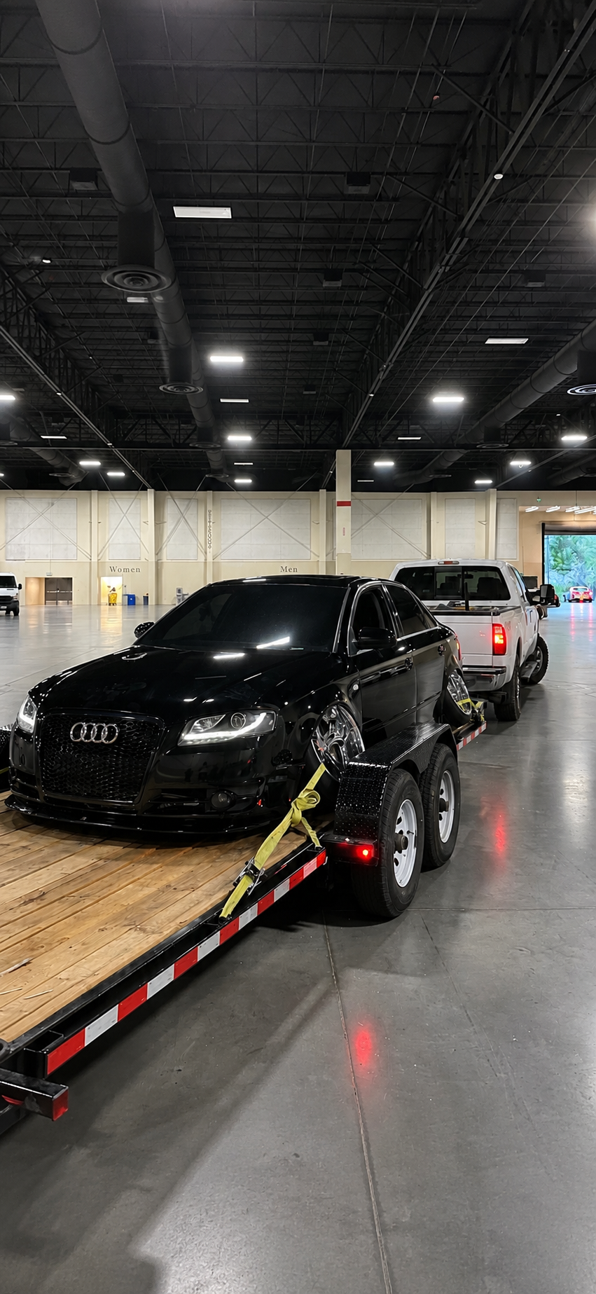 Black Audi sedan strapped on a wooden trailer hitched to a white pickup truck in a large garage.
