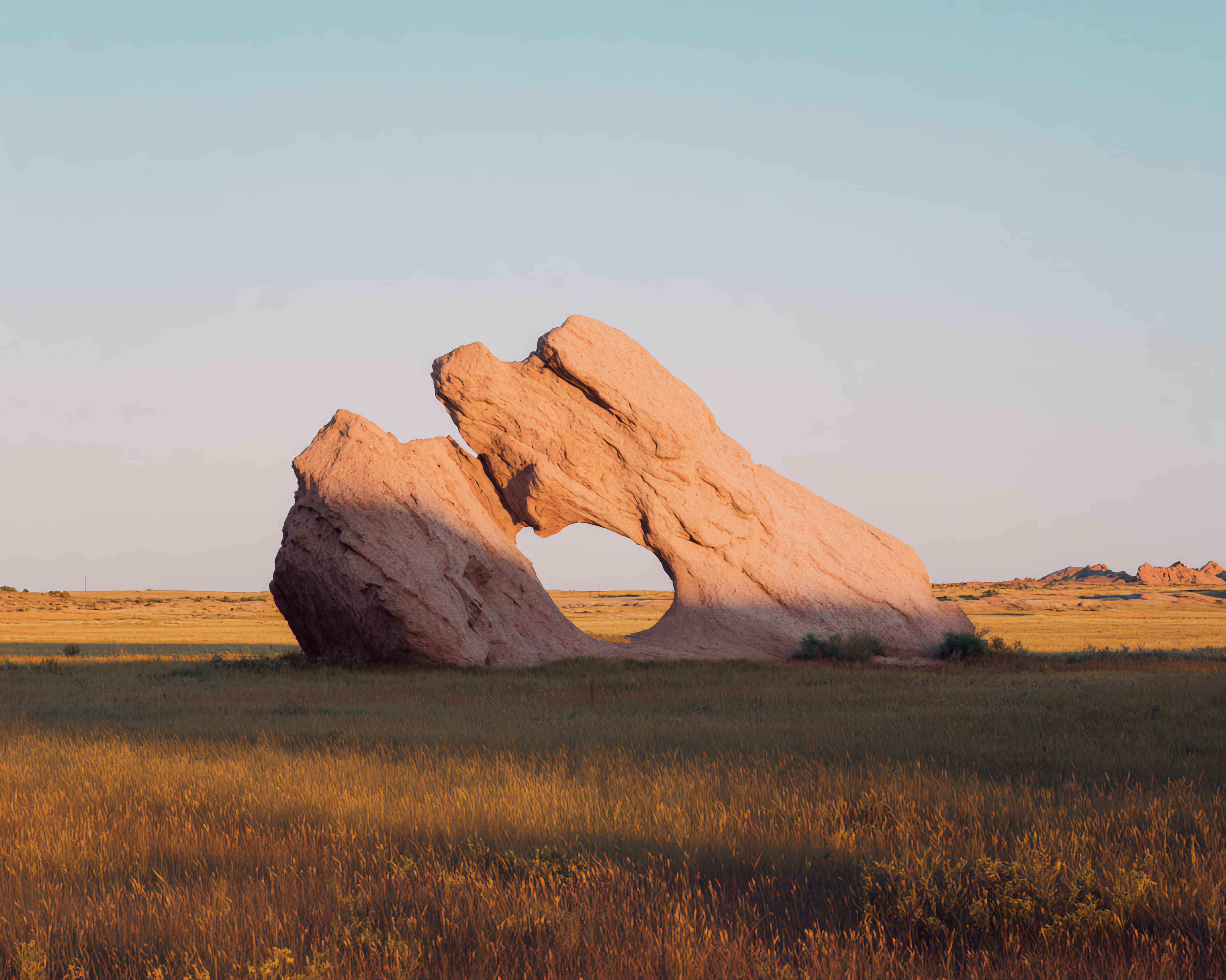 Large pink rock formation with a hole in the center stands in a grassy field under clear sky.