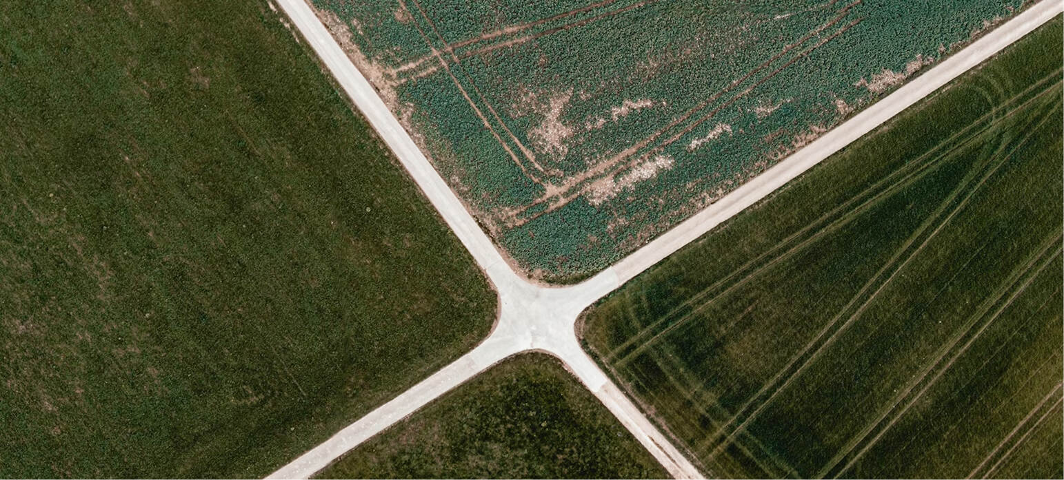 Aerial view of a dirt crossroads dividing green agricultural fields with crop patterns.