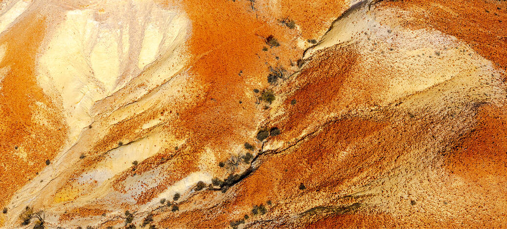 Aerial view of dry, rugged landscape with red and beige soil and sparse vegetation.