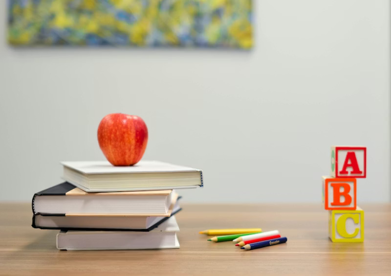 Red apple on top of a stack of books with colored pencils and ABC blocks on a wooden surface.
