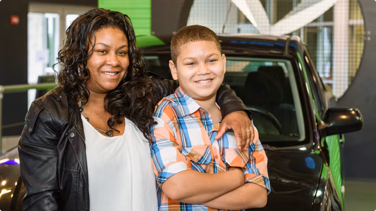 Smiling woman with arm around a boy in plaid shirt standing in front of a black car indoors.