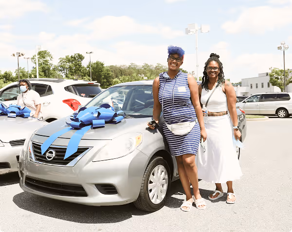 Two smiling women stand beside a silver car with a large blue bow in a sunny parking lot.