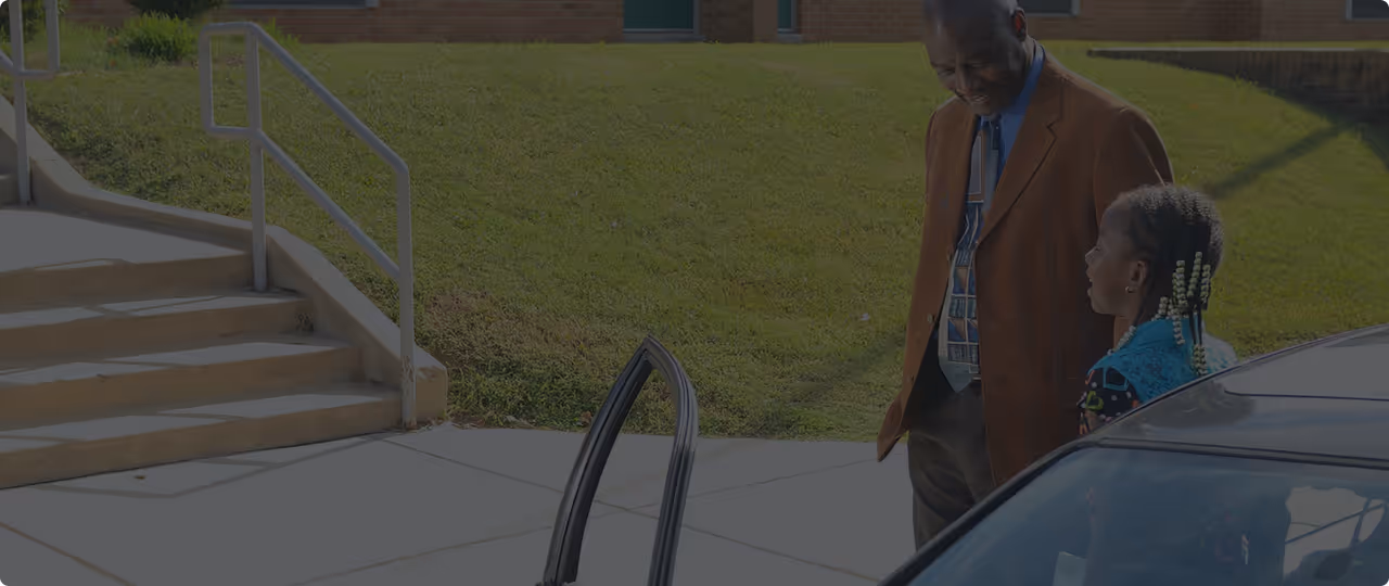 Man in brown jacket talking to young girl with braided hair beside a car outdoors.