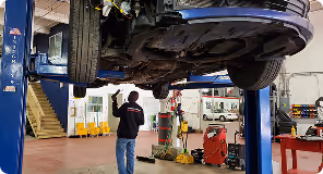 Car lifted on a hydraulic hoist in a workshop with a mechanic standing underneath.