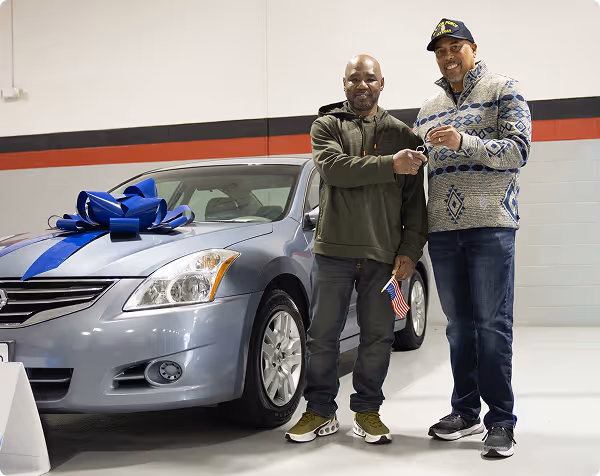 Two men shaking hands beside a silver car with a large blue bow in a garage.