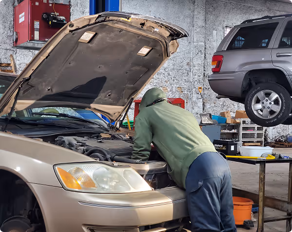 Mechanic in a green hoodie inspecting the engine of a beige car with its hood open in a garage.