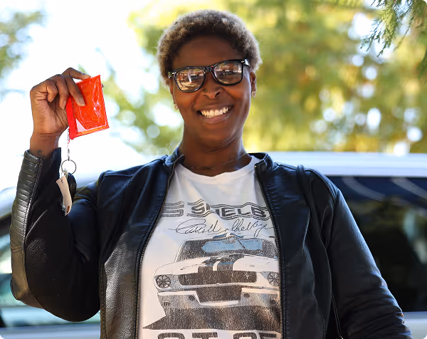 Smiling woman with glasses, holding a red wallet and car keys outdoors near a vehicle.