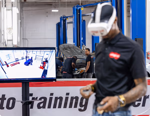 Man using virtual reality headset in automotive training center while others work on a car in background.