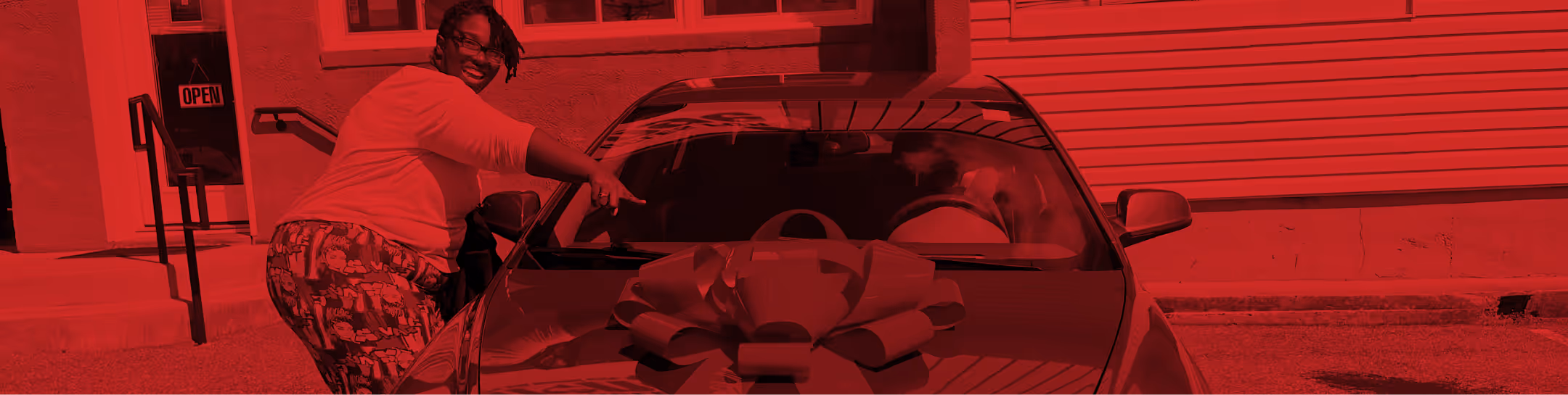 Person smiling and pointing at a new car with a large bow on its hood outside a building.