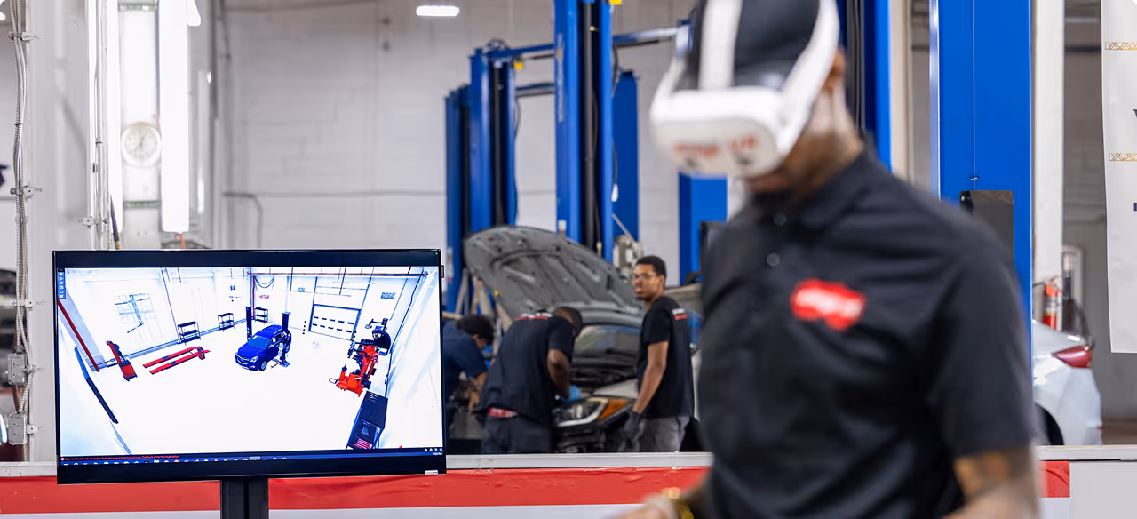 Man wearing a VR headset in an auto repair shop with a monitor showing a virtual garage scene.