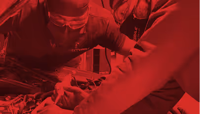 Two mechanics working together under a car hood in a garage, wearing gloves and safety glasses.