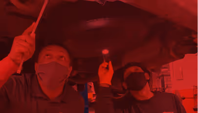 Two masked mechanics inspecting a car underside using a flashlight and tool in a garage.