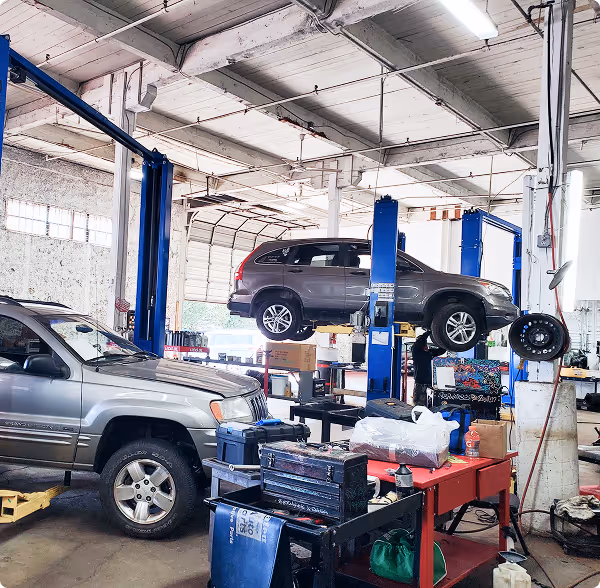 Car repair shop with an SUV lifted on a blue hydraulic lift and another vehicle parked inside.