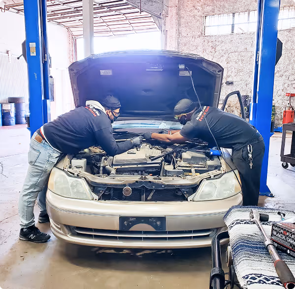 Two masked mechanics working on a car engine under an open hood in a workshop.