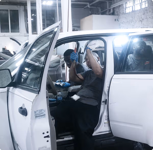 Two mechanics working inside and beside a white car in a garage with open doors.