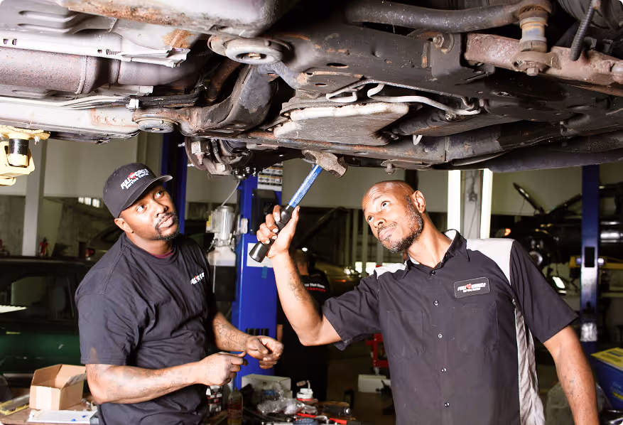 Two mechanics inspecting and repairing the underside of a vehicle in a garage.