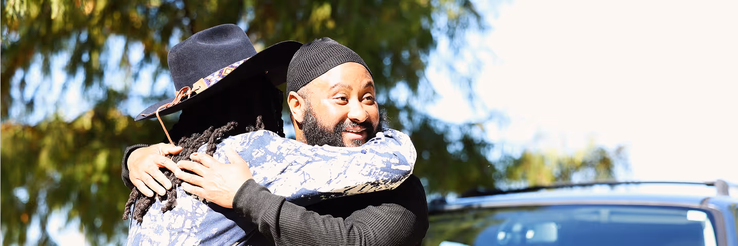 Two men smiling and hugging outdoors near a car and trees on a sunny day.