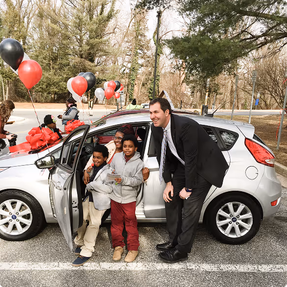 Man in suit with three smiling children standing beside a silver car decorated with red and black balloons.