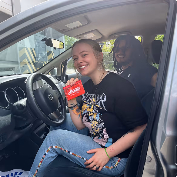 Smiling woman holding a red card seated in the driver seat of a parked car with a man in the back seat.