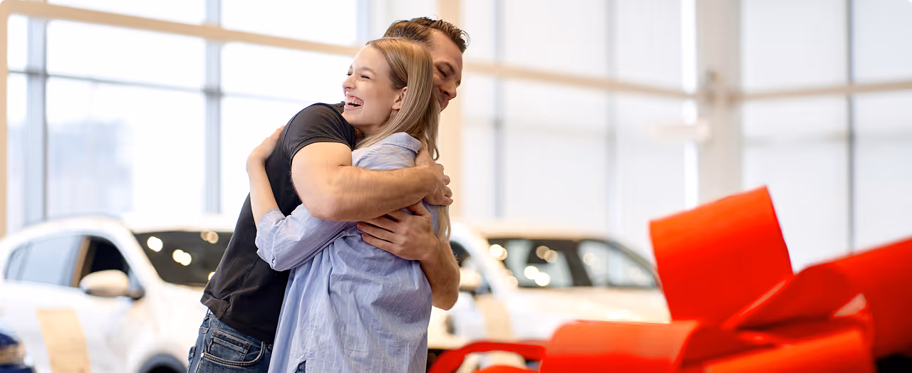 Smiling couple hugging happily in bright showroom with white cars and a large red bow.