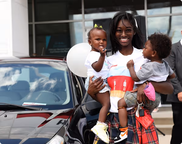 Smiling woman holding two toddlers in front of a black car outside a building.