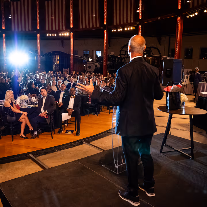 Man in black suit speaking to a seated audience at a formal indoor event with stage lighting.