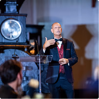 Man in tuxedo giving a speech at a podium with a vintage train in the background.