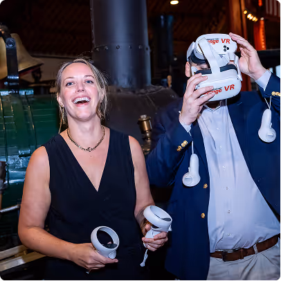 Woman smiling holding VR controllers and man wearing a VR headset indoors at an event.
