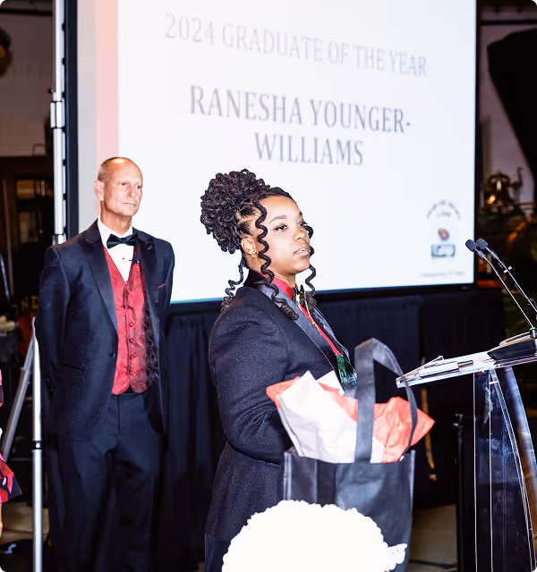 Woman stands at podium receiving 2024 Graduate of the Year award with man in tuxedo behind her.