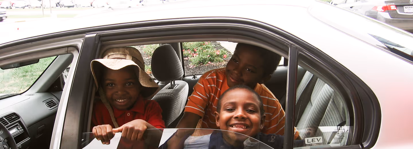 Three smiling children sit inside a car, looking out of the open window.