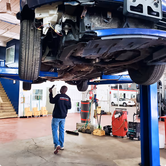 Mechanic walking under a car lifted on a blue hydraulic lift in an auto workshop.