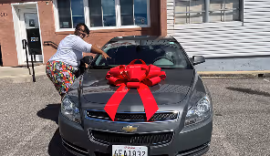 Woman standing beside a gray Chevrolet car with a large red bow on the hood in a parking lot.