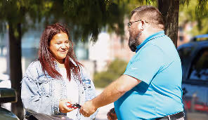 Man in blue shirt handing car keys to smiling woman with red hair outdoors.