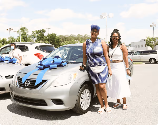 Two smiling women standing beside a silver Nissan car with a large blue bow on the hood in a parking lot.