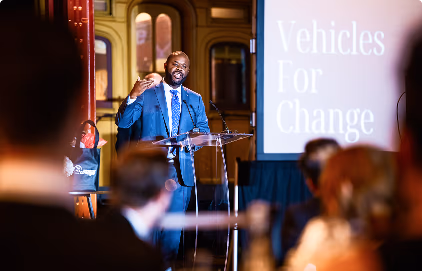 Man in a suit speaking at a clear podium to an audience with 'Vehicles For Change' displayed behind him.