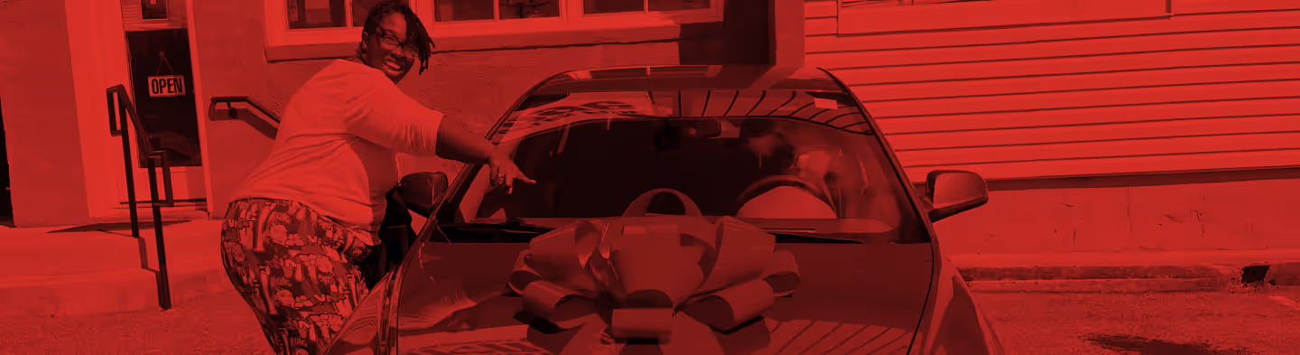 Woman smiling while pointing at a car with a large bow on its hood parked outside a building.