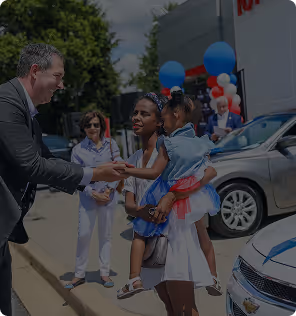 Man in suit shaking hand with woman holding a child near parked cars and balloons outdoors.