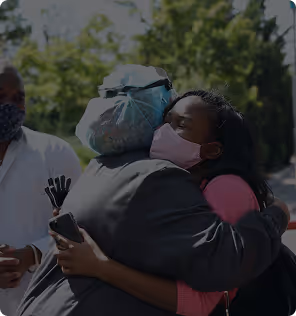 Two women wearing masks embrace outdoors while a man in a mask stands nearby.