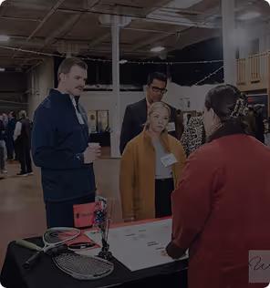People gathered around a table with tennis rackets and a red box at an indoor event.
