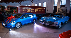 Blue modern hatchback with a red bow beside a vintage blue convertible inside a showroom.