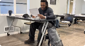 Man sitting at a classroom desk reading a paper with bags beside him and empty desks nearby.