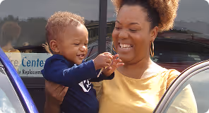 Smiling woman holding and playing with a happy toddler near a car.