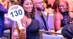 Smiling woman holding auction paddle number 130 at a seated event with other attendees in background.